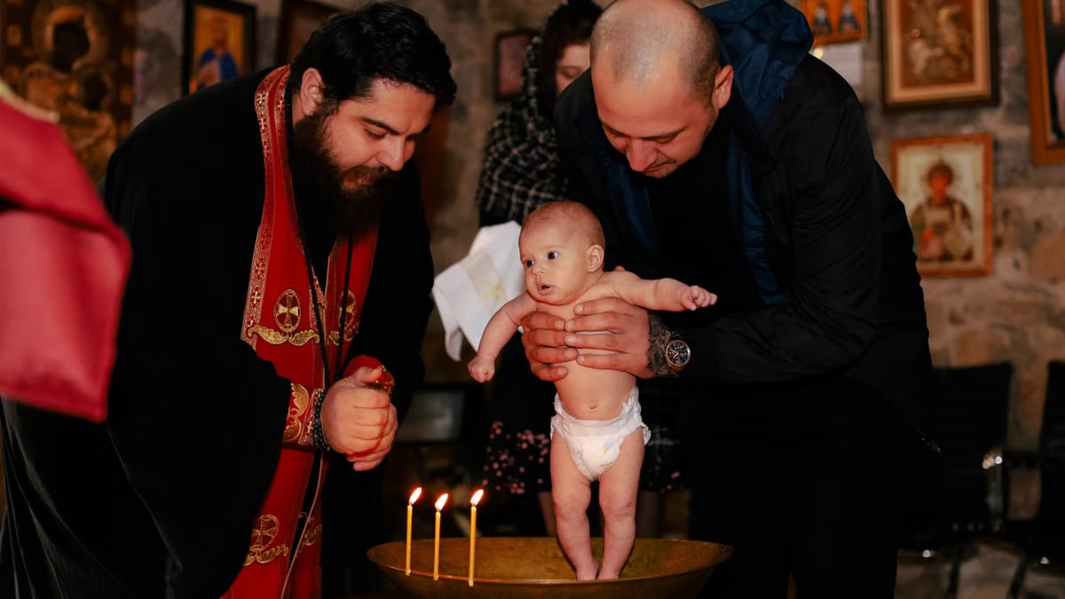 A priest and father performing an Orthodox baptism ceremony with a baby.